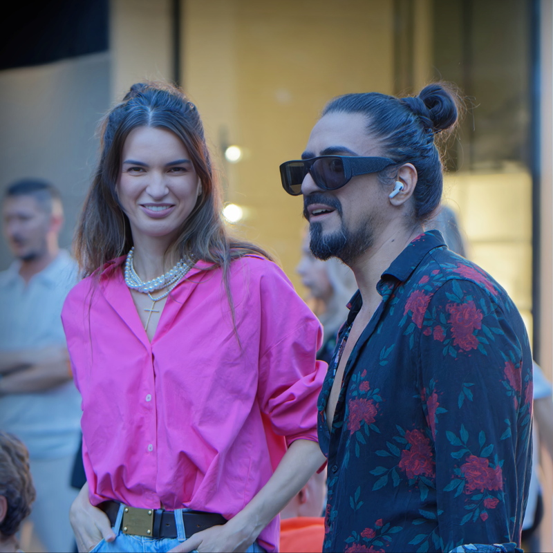 14 - A woman in a bright pink button-down shirt layered with pearl necklaces and a man in a floral-print black shirt with dark sunglasses and his hair in a bun